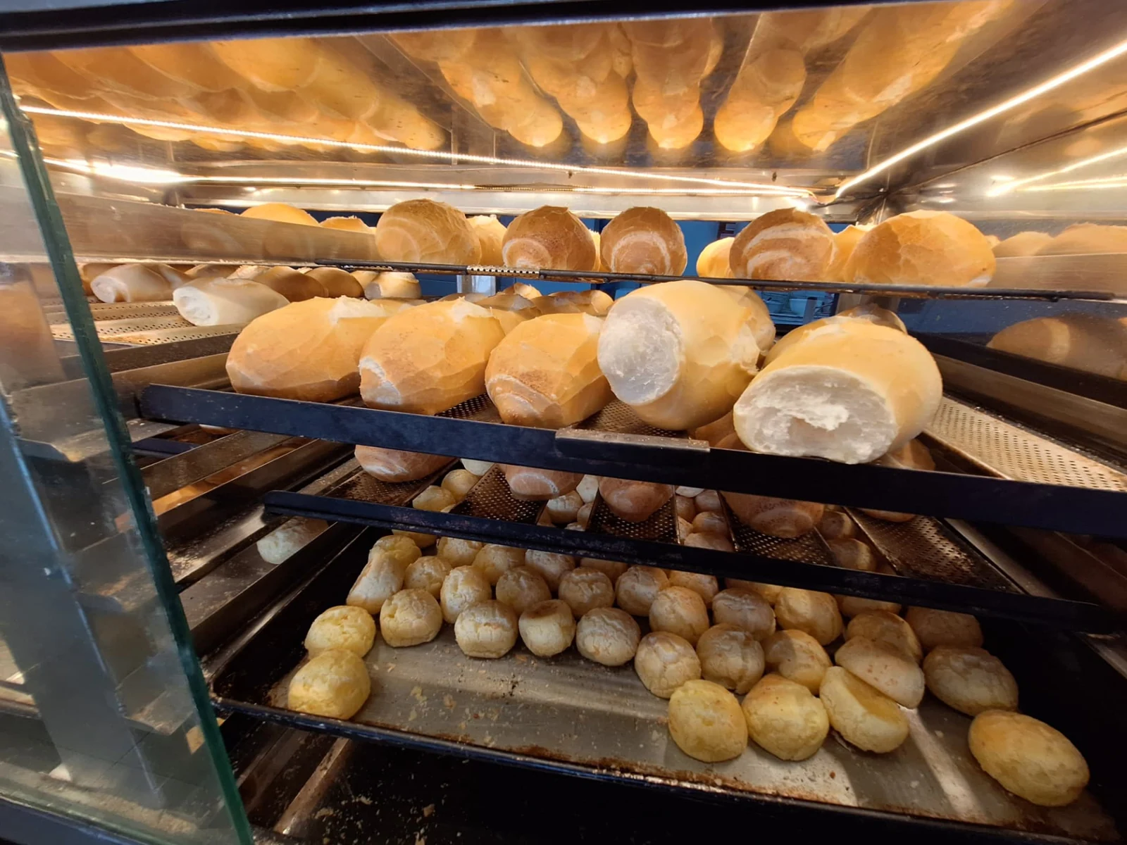 Pães franceses dourados e pães de queijo em bandejas na vitrine iluminada da ManáPan