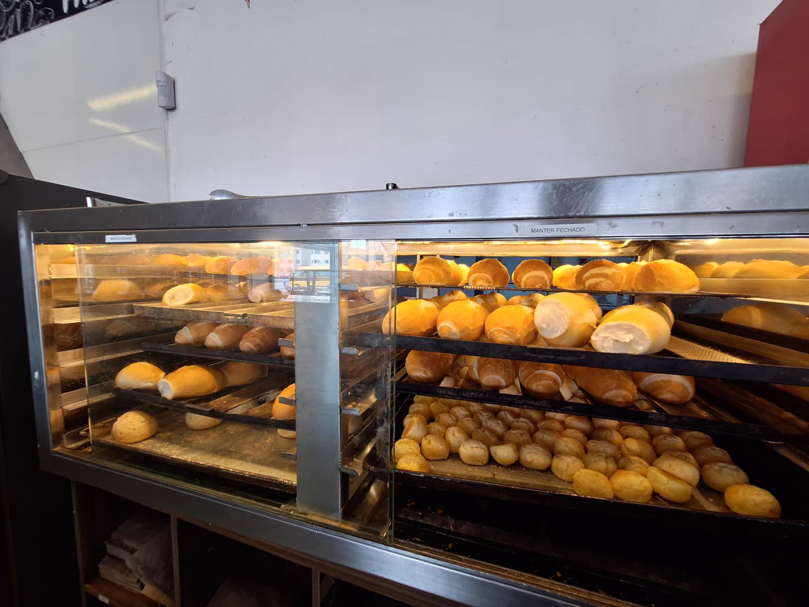 Estufa aquecida da ManáPan com pães franceses dourados nas prateleiras e bandeja de pão de queijo na parte inferior