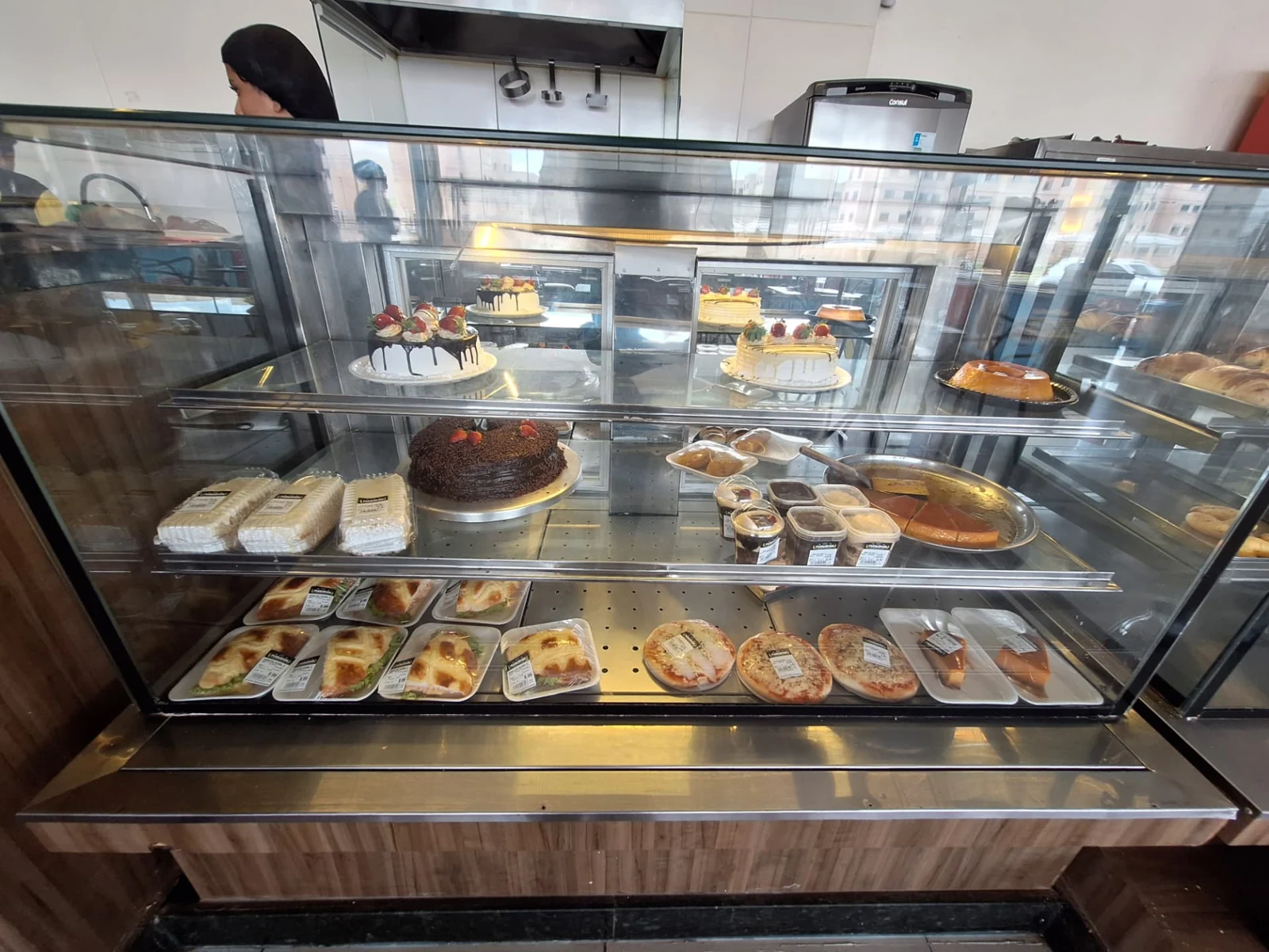 Vitrine refrigerada da ManáPan com tortas decoradas com morangos, bolo de chocolate, pudins e sobremesas em potes